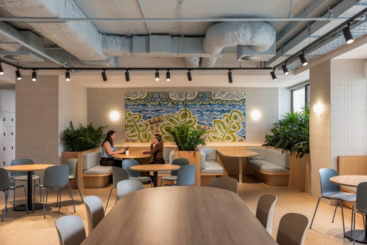 An office kitchen with a large feature artwork on the back wall. The artwork has green leaves and blue swirls to represent the ocean. There are two women sitting in front of the artwork at a booth, chatting.